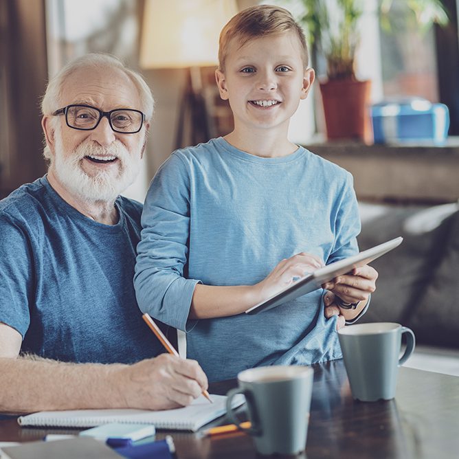 Positive delighted boy holding tablet in both hands Have a rest. Cheerful kid looking straight at camera while standing near his grandfather